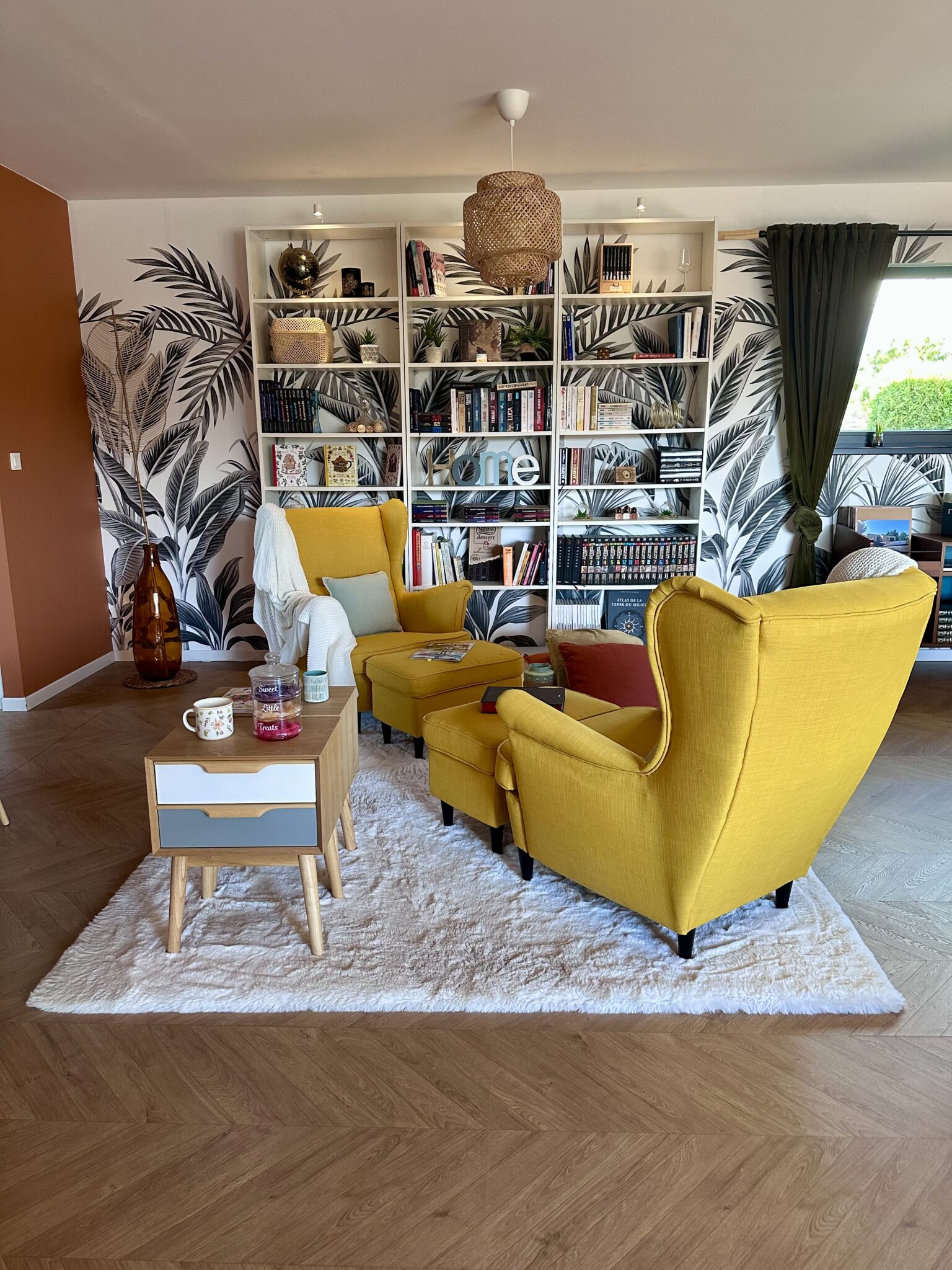 Coin lecture chaleureux avec fauteuils jaunes, bibliothèque sur mesure, papier peint végétal et parquet en chevrons.
